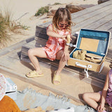 A child applying sun cream sitting next to an open olli ella see-ya suitcase filled with things including a matching butterscotch coloured wash bag