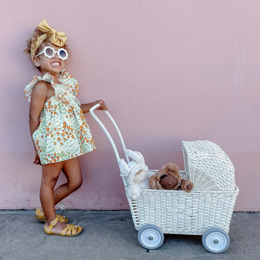 Girl stood in front of a pink wall pushing the Olli Ella chalk coloured rattan strolley toy