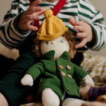 Toddler dressing Charlie Chestnut, a Dress Me Dinky Doll by Olli Ella, complete with a removable cosy green fleece trench coat, yellow bobble beanie hat.