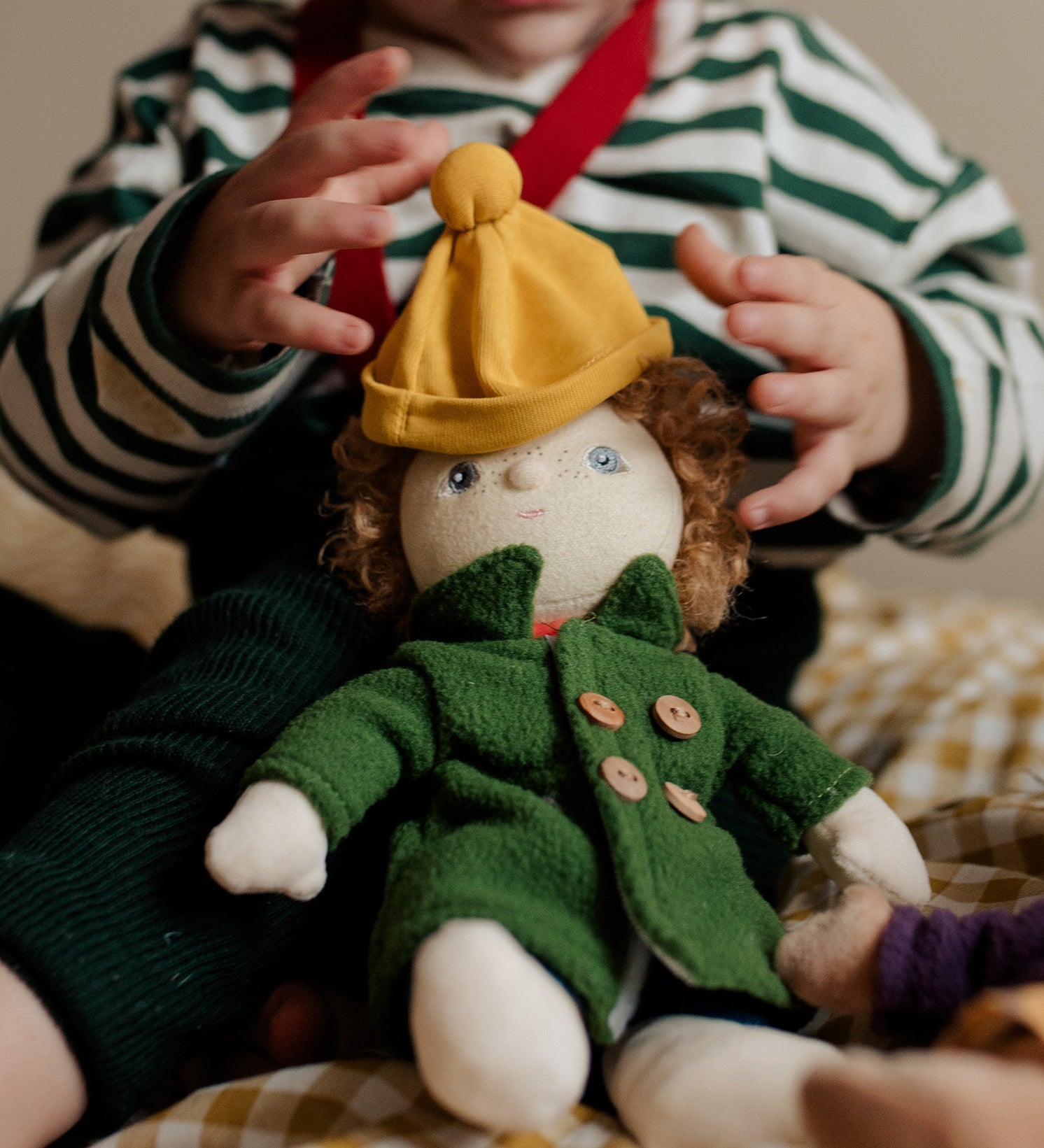 Toddler dressing Charlie Chestnut, a Dress Me Dinky Doll by Olli Ella, complete with a removable cosy green fleece trench coat, yellow bobble beanie hat.