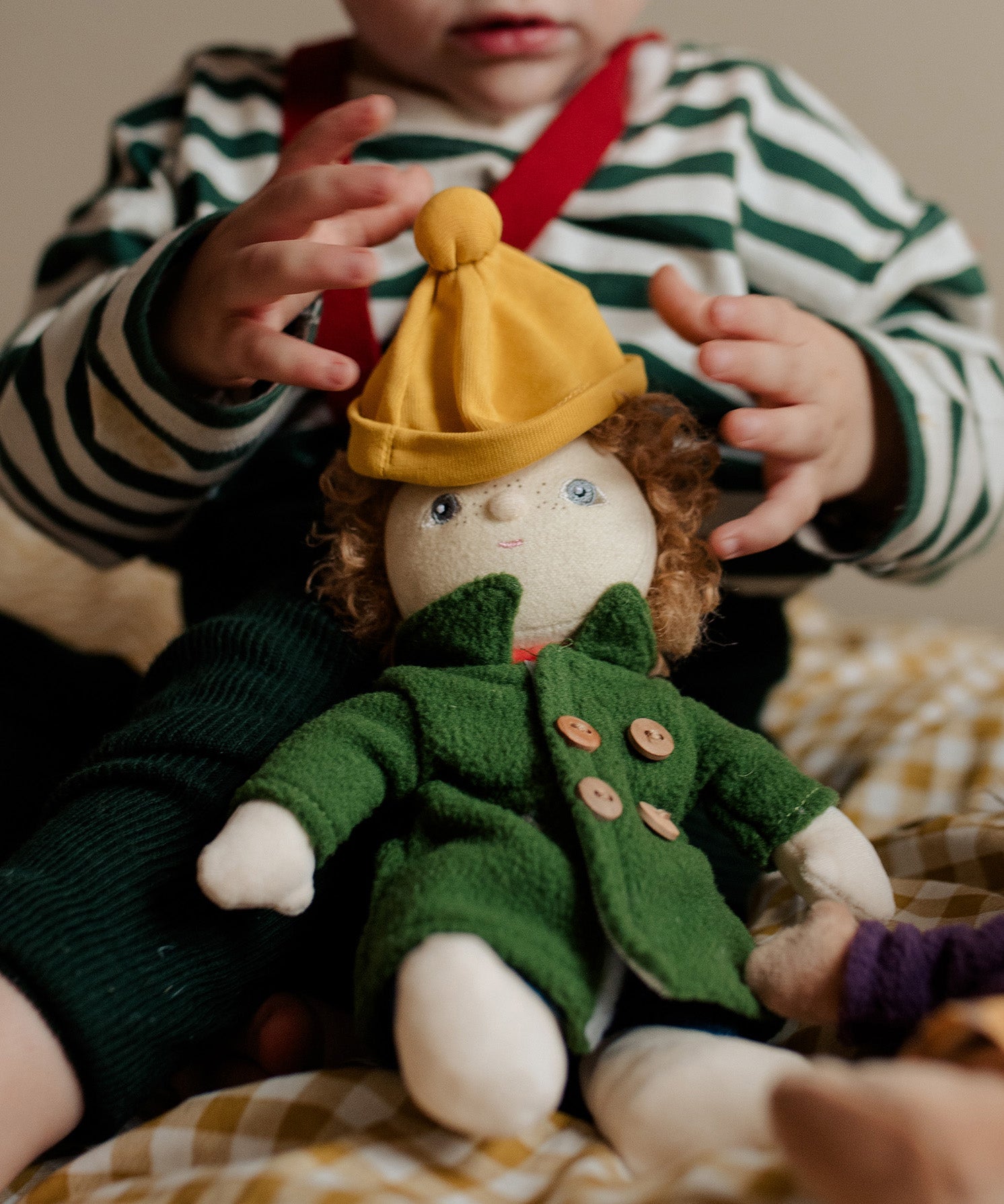 Toddler dressing Charlie Chestnut, a Dress Me Dinky Doll by Olli Ella, complete with a removable cosy green fleece trench coat, yellow bobble beanie hat.