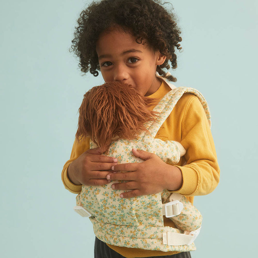 Close up of young child holding an Olli Ella dinkum doll in a floral print childrens toy carrier on a blue background