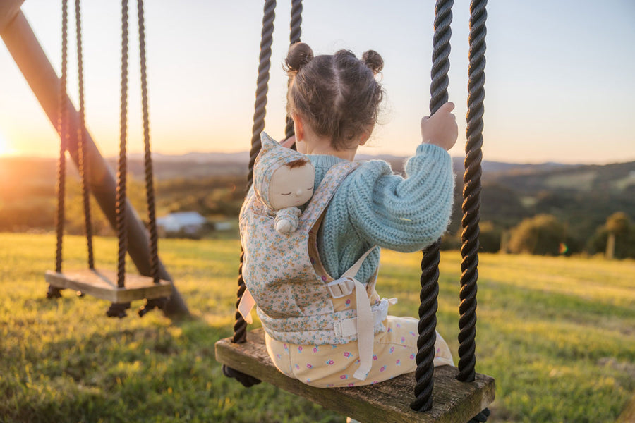 Young girl sat on a swing wearing the Olli Ella floral print dozy dinkum doll baby carrier on her back