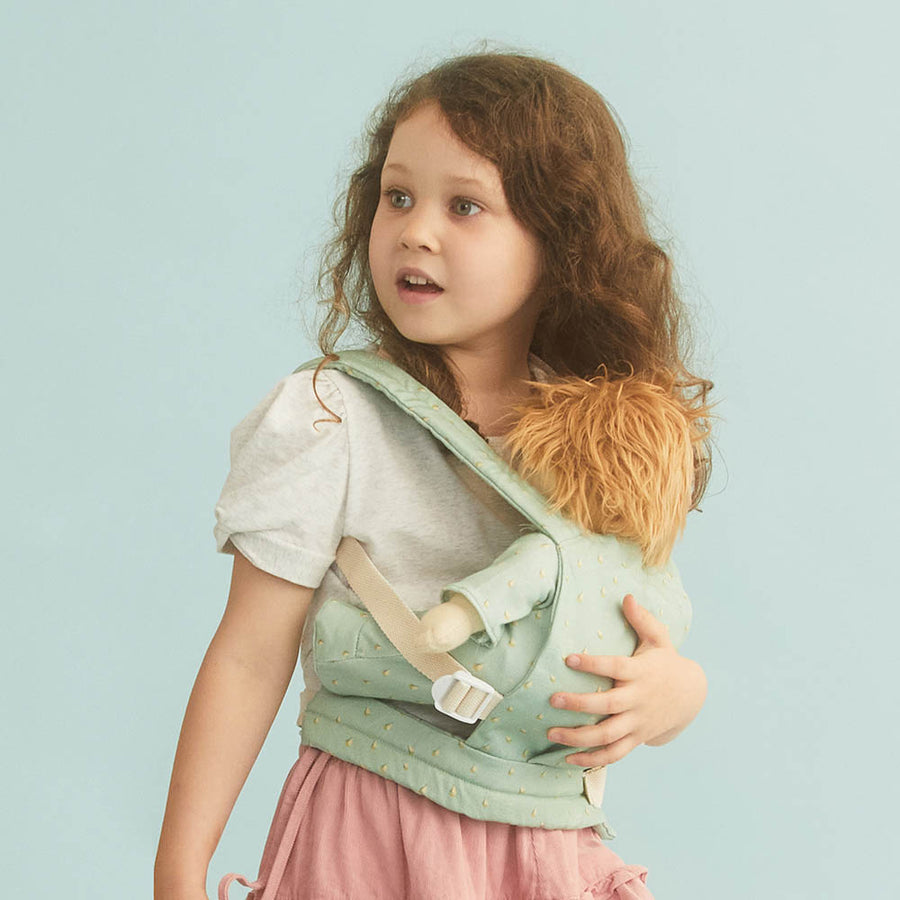 Young girl stood on a blue background carrying an Olli Ella dinkum doll in the boat print doll baby carrier