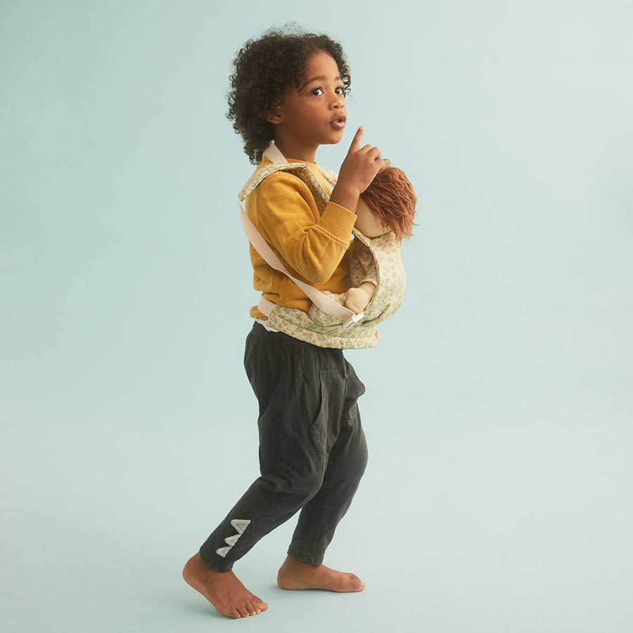 Young child stood on a blue background carrying an Olli Ella dinkum doll in the blossom print toy doll baby carrier