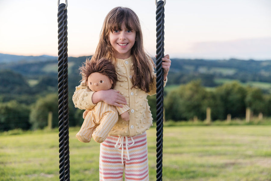 Young girl stood next to a rope swing holding an Olli Ella dinkum doll dressed in the goldie striped pyjamas