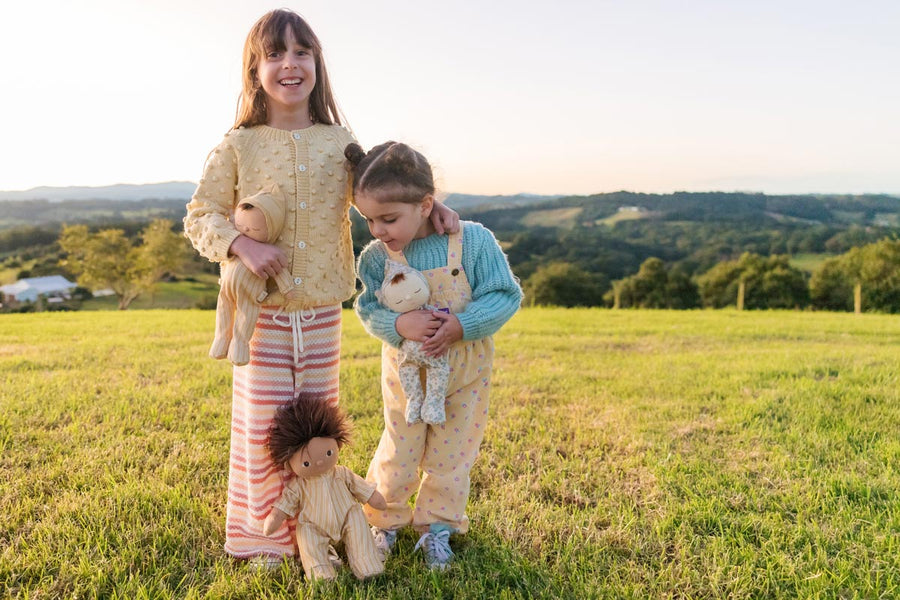 Two young girls stood in a grass field holding some Olli Ella dozy dinkum doll toys dressed in the goldie stripe pyjamas