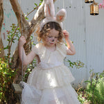 A child wearing the Olli Ella Fluffle Bunny Ear Headband in Pink. The child is wearing a cream coloured dress sitting on a swing. 