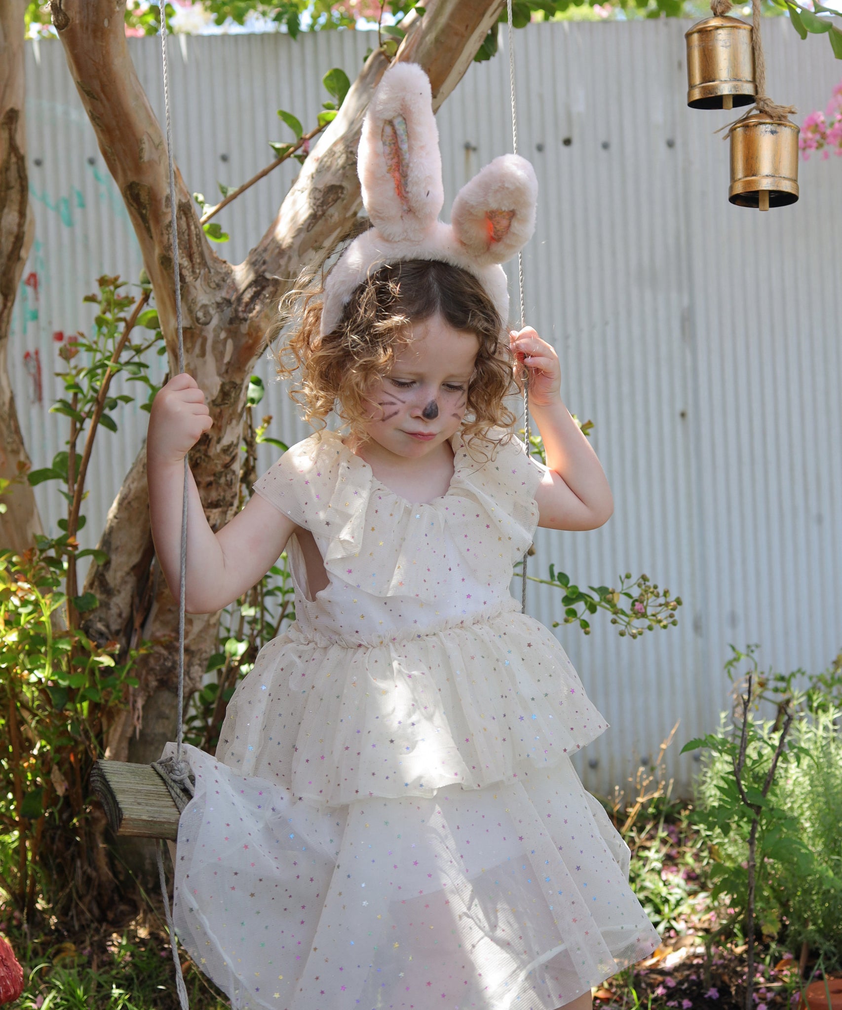 A child wearing the Olli Ella Fluffle Bunny Ear Headband in Pink. The child is wearing a cream coloured dress sitting on a swing. 