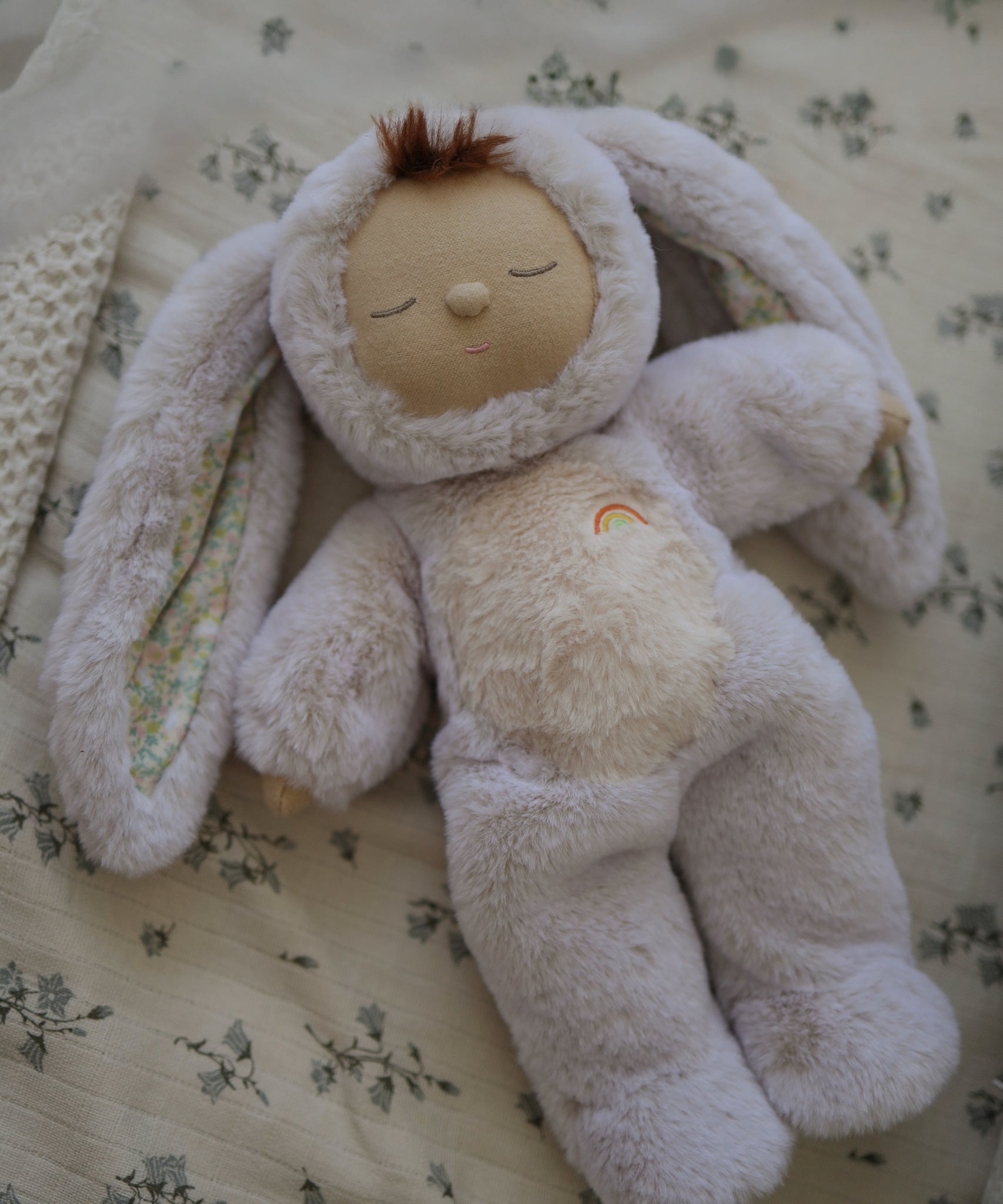 The Olli Ella Bunny Lala Cozy Dinkum soft toy laying on a floral patterned blanket. 