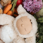 A close up of the Lamby Picket Olli Ella Cozy Dinkum Doll laying besides flowers and vegetables. 