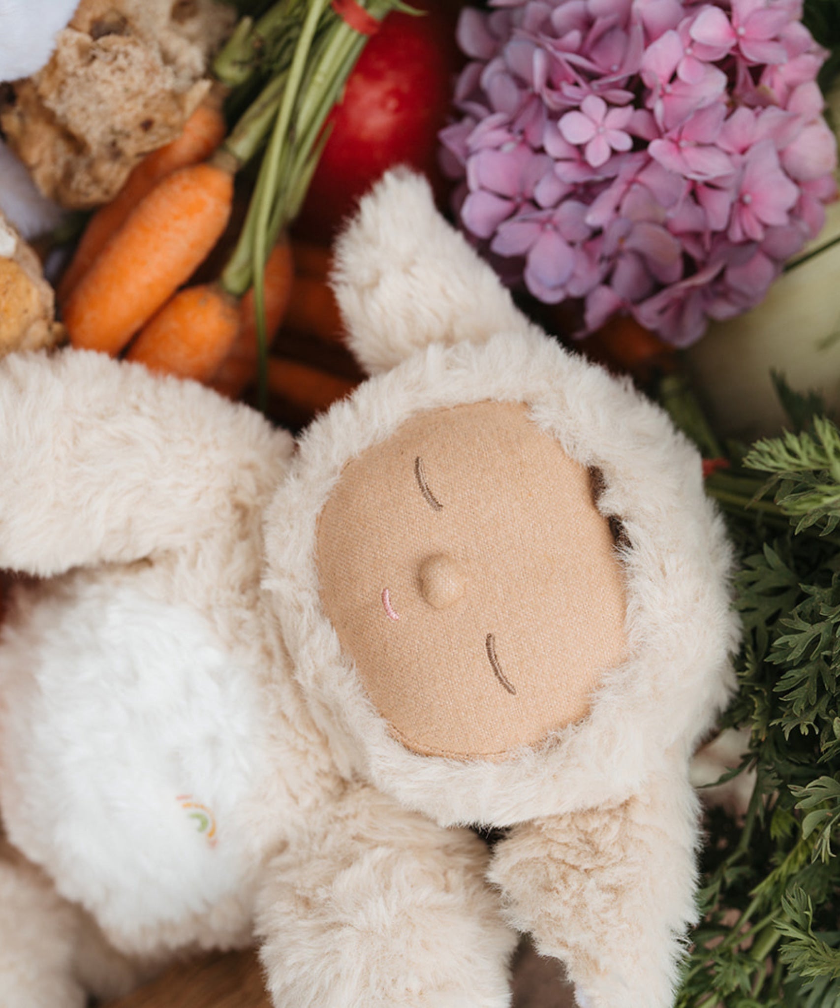 A close up of the Lamby Picket Olli Ella Cozy Dinkum Doll laying besides flowers and vegetables. 