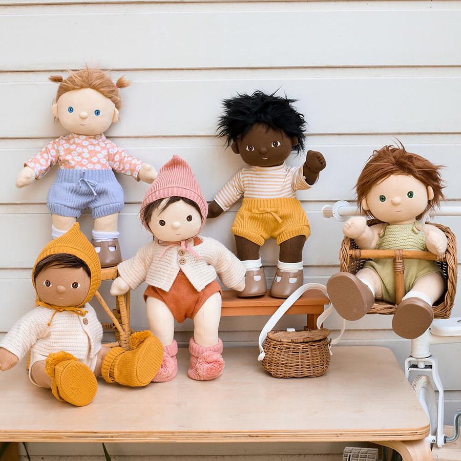 5 Olli Ella dinkum dolls dressed up in accessory clothing on a wooden table in front of a white fence