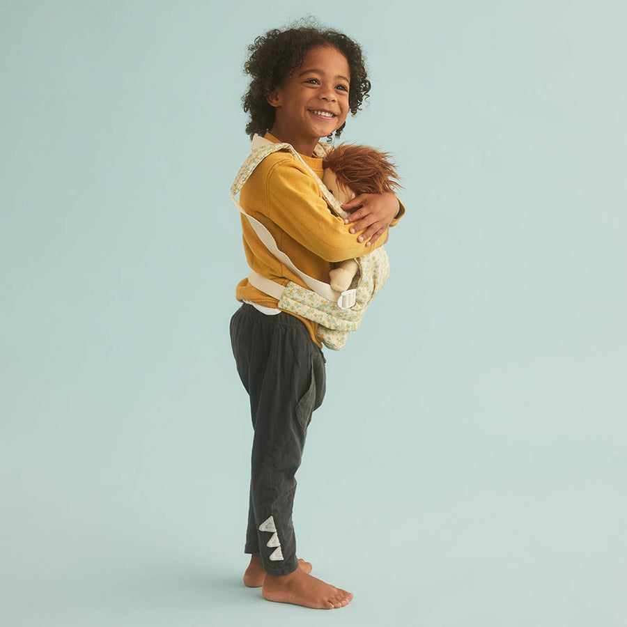 Young child stood on a blue background carrying an Olli Ella dinkum doll in the floral print baby toy carrier