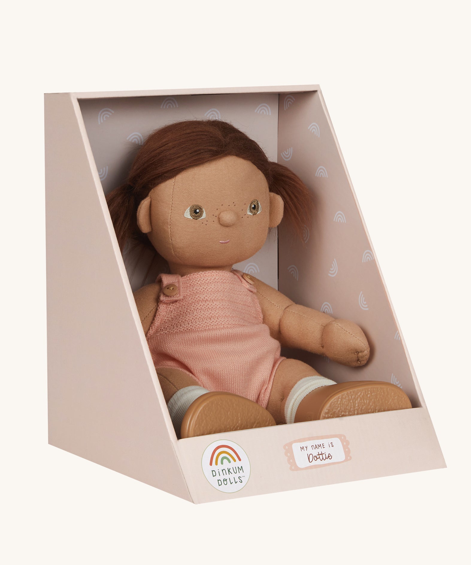 An Olli Ella Dottie Dinkum Doll on a plain background shown in a seated position in it's box. The doll has brown hair and brown eyes. The hair is in pig tails and the doll is a seated position with just the nappy on. 