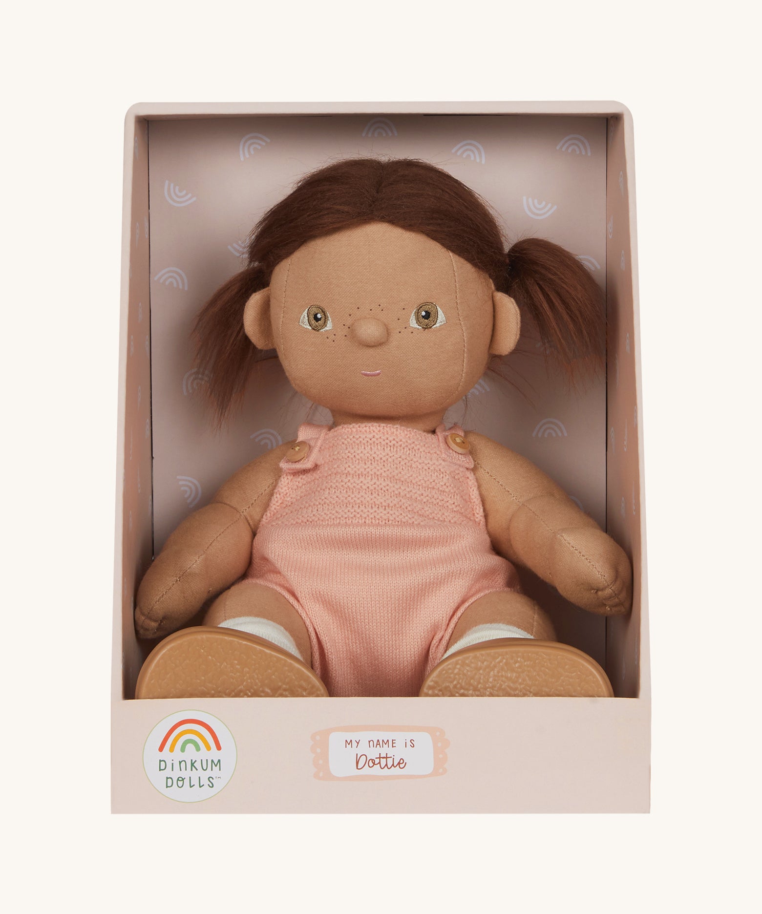 An Olli Ella Dottie Dinkum Doll on a plain background shown in a seated position in it's box. The doll has brown hair and brown eyes. The hair is in pig tails and the doll is a seated position with just the nappy on. 