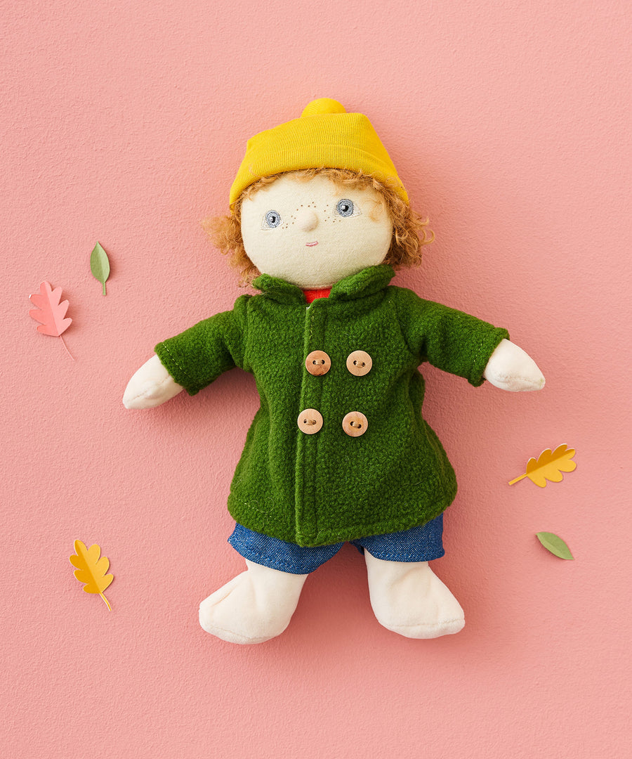 Charlie Chestnut is a Dress Me Dinky Doll by Olli Ella, complete with a removable cosy green fleece trench coat, yellow bobble beanie hat and denim shorts . Laying flat on a pink background surrounded by paper leaves.