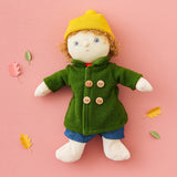 Charlie Chestnut is a Dress Me Dinky Doll by Olli Ella, complete with a removable cosy green fleece trench coat, yellow bobble beanie hat and denim shorts . Laying flat on a pink background surrounded by paper leaves.