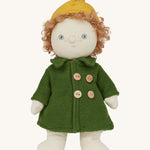 Charlie Chestnut is a Dress Me Dinky Doll by Olli Ella, complete with a removable cosy green fleece trench coat, yellow bobble beanie hat . Sat down, cream background.