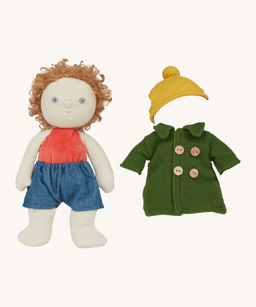 Charlie Chestnut Dress Me Dinky Doll by Olli Ella, wearing denim shorts. The green fleece trench coat, and yellow bobble beanie hat next to him. Cream background. 