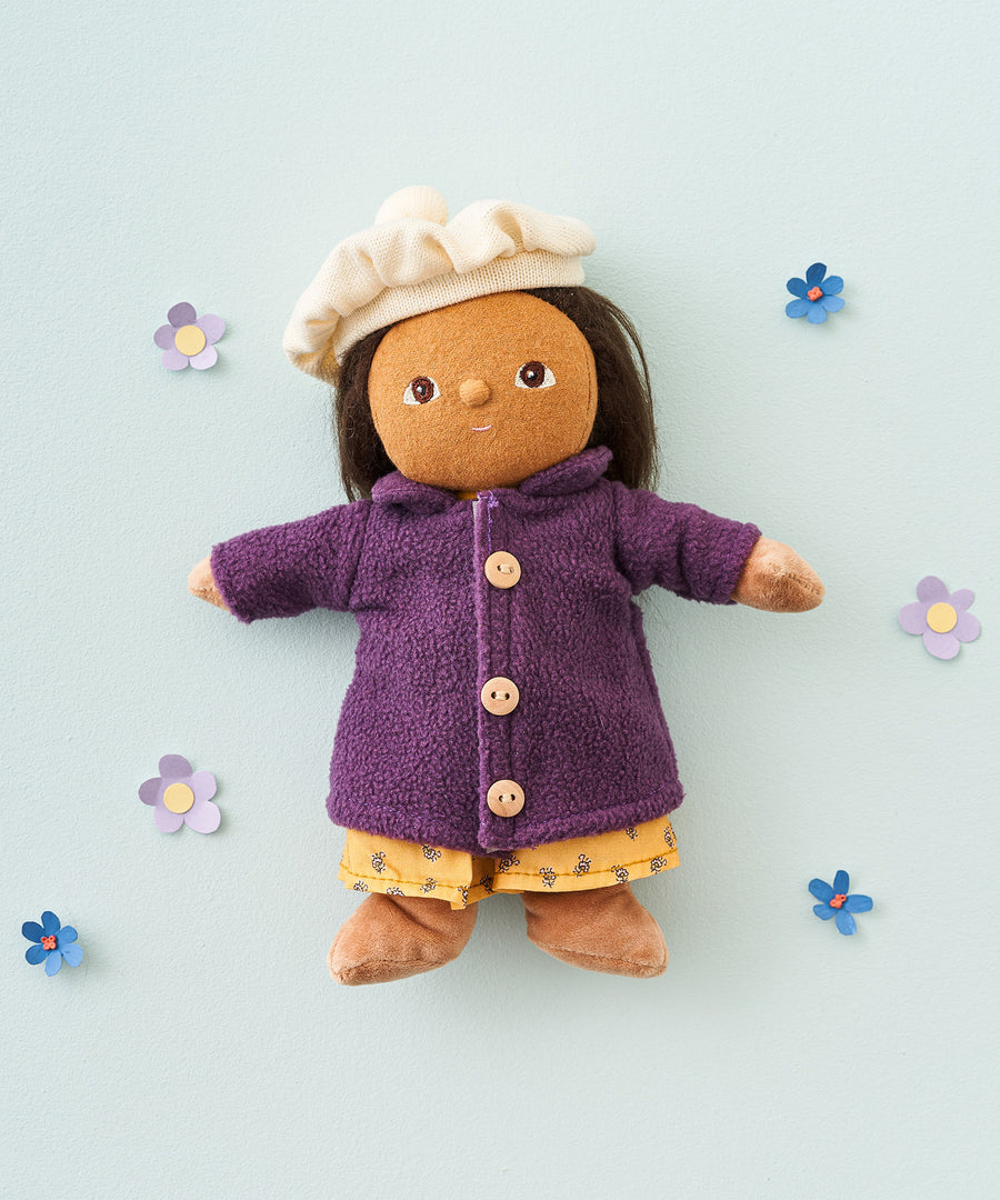 Olli Ella Dress Me Dinky - Lila Lavender wearing a purple fleece trench coat, yellow dress and white beret. Surrounded by paper flowers, blue background. 