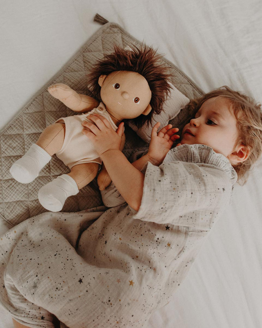 Girl laying on a bed with her arm around an Olli Ella dinkum doll on a soft seafoam strolley bedding set