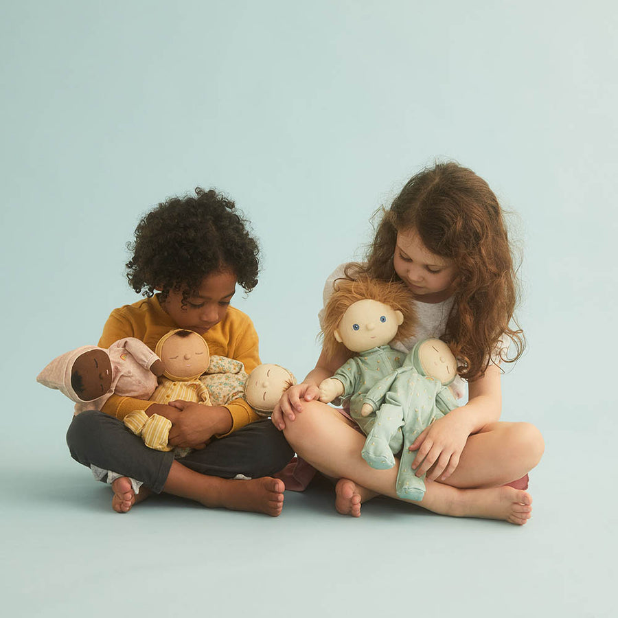 Two young children sat cross legged playing with some Olli Ella dozy dinkum dolls on a blue background