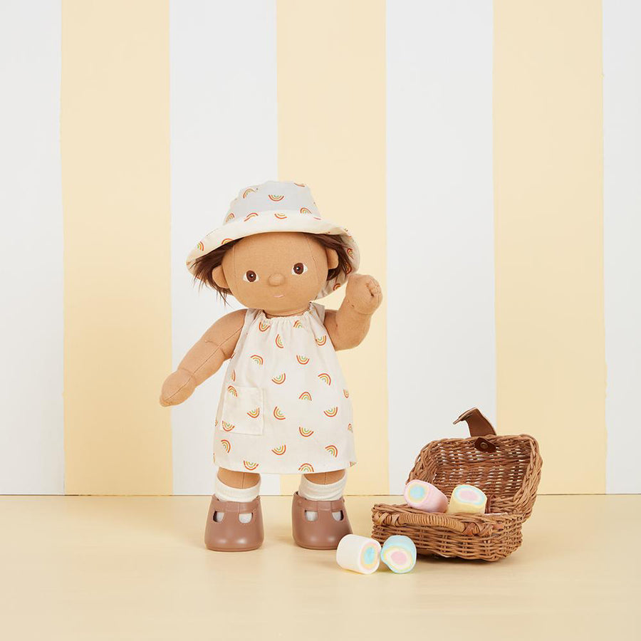 Olli Ella dinkum doll stood on a cream background next to a wooden basket wearing the una rainbow dress set