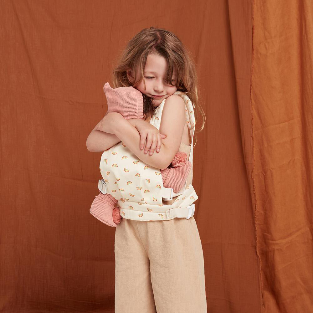 Girl cuddling a dinkum doll toy in her Olli Ella soft rainbow baby carrier accessory in front of a brown sheet background