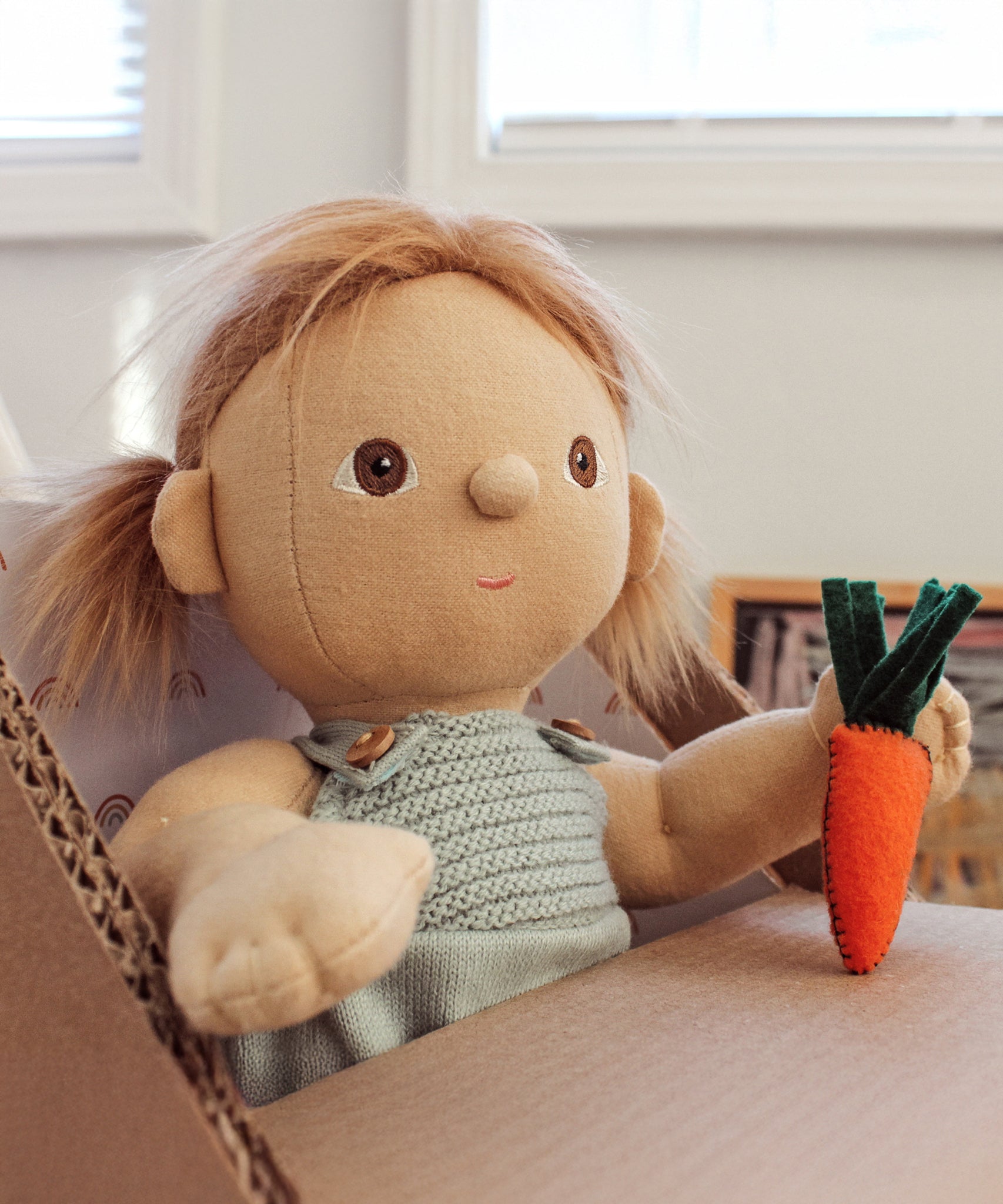 An Olli Ella Gigi Dinkum doll holding a felt carrot sitting in a home made cardboard high chair. 