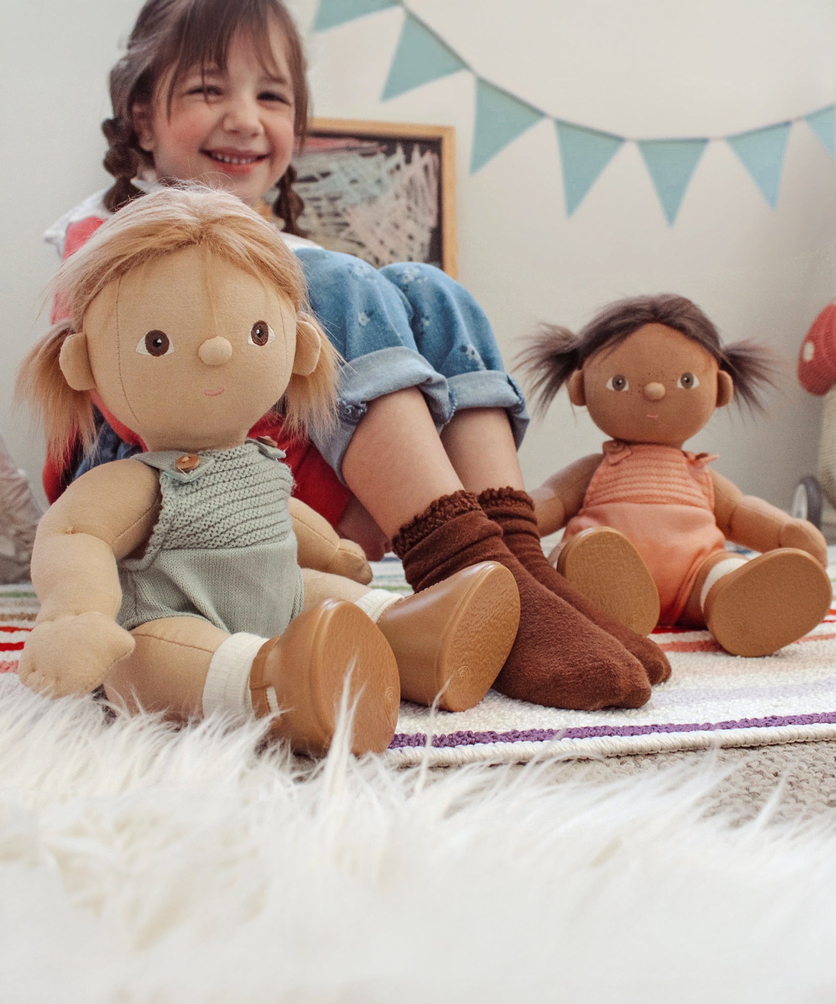 An Olli Ella Gigi Dinkum doll sat on striped rug, the Dottie doll can be seen in the background sitting next to a child. 