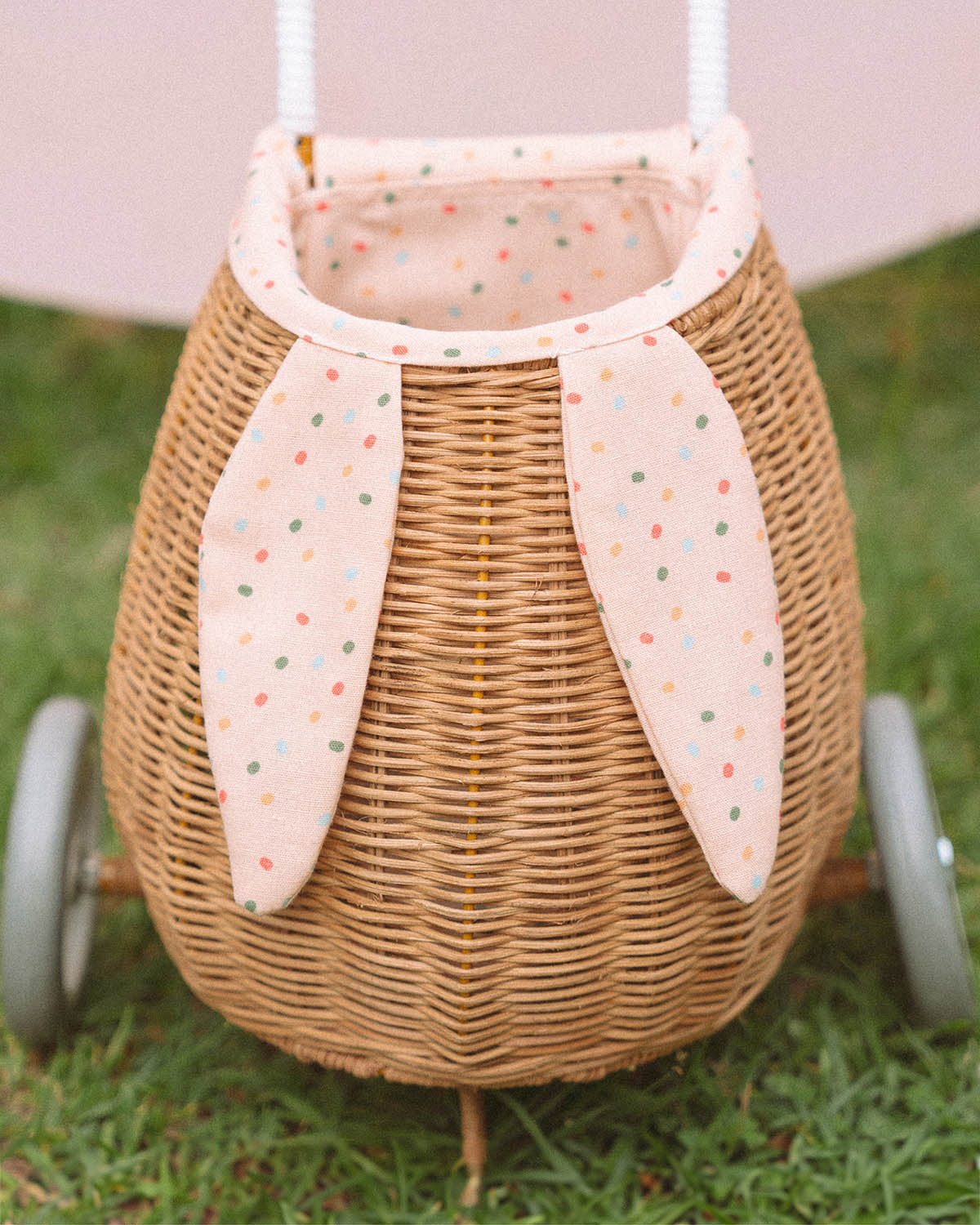 A child happily stood in the sun next to the Olli Ella Rattan Bunny Luggy with Lining – Gumdrop and holding the Rattan basket in Gumdrop