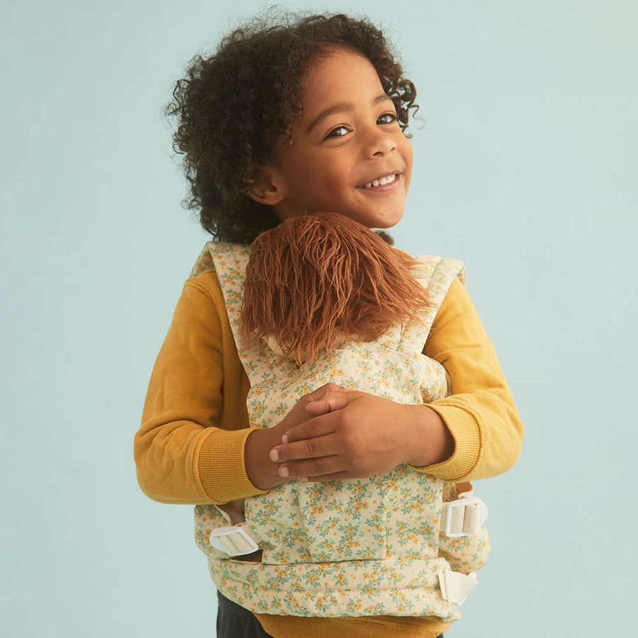 Close up of young child hugging an Olli Ella dinkum doll in the floral print baby doll carrier on a blue background