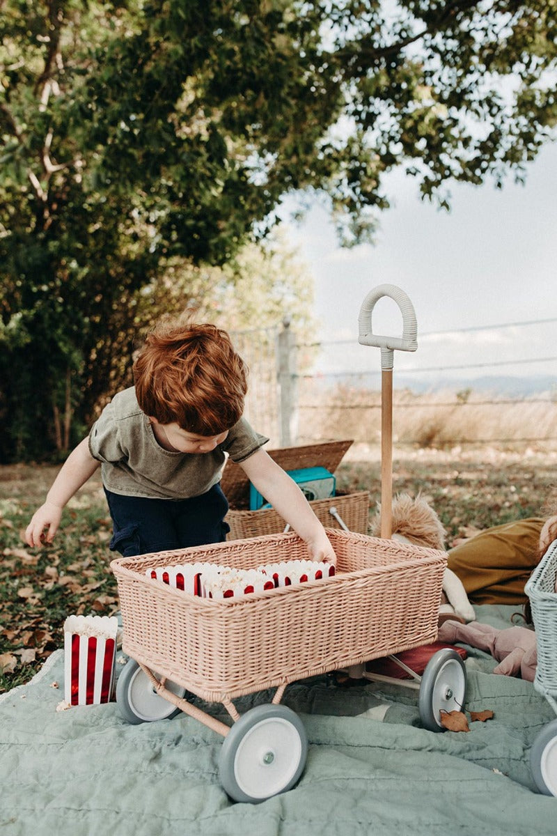 Boy crouched down on a blue blanket pulling popcorn from inside the Olli Ella pink rattan wonder wagon