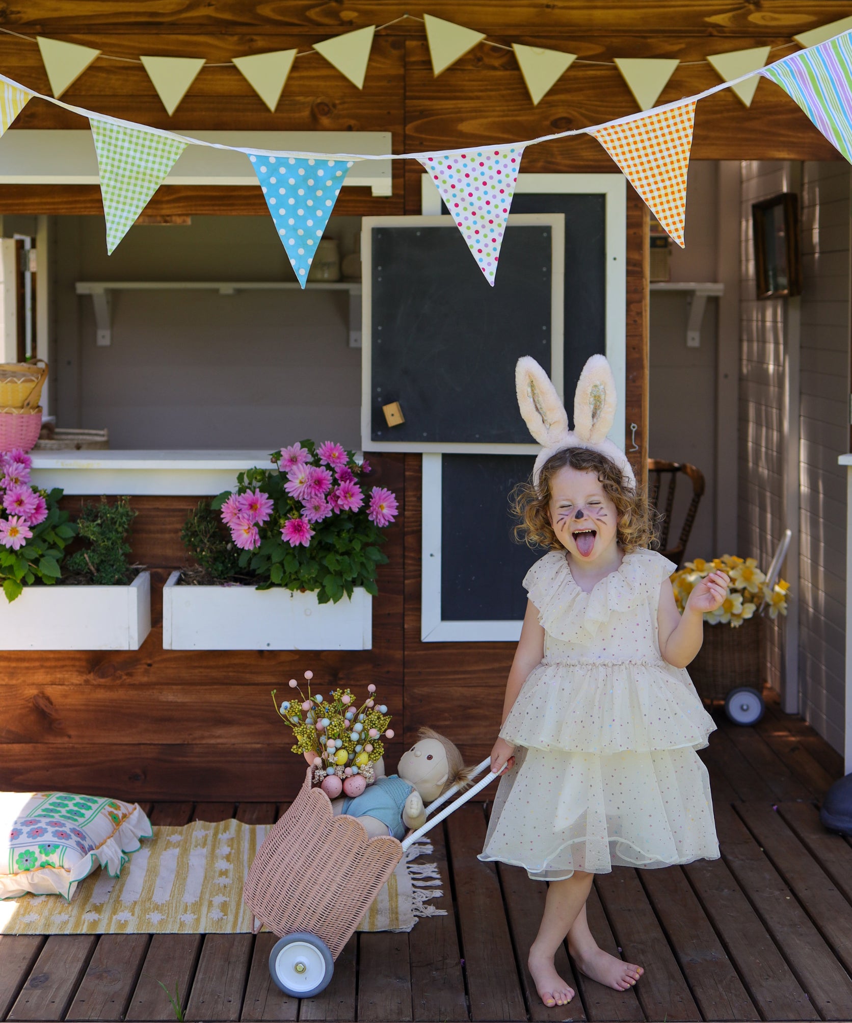 A child wearing the pink Olli Ella Fluffle Bunny Ear Headband. The child is dressed in a cream coloured dress pulling along an olli ella dinkum doll in a luggy style basket. 