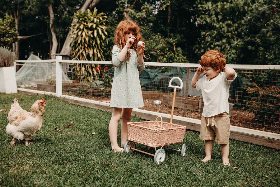 Girl and boy stood on some grass next to the Olli Ella light pink rattan wonder wagon toy