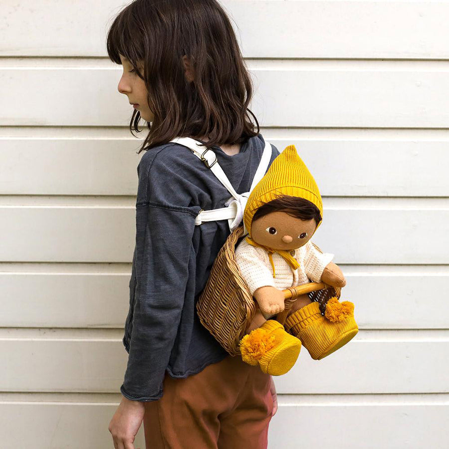 Girl stood in front of a white fence wearing a wooden backet backpack with a dinkum doll sat inside wearing the honey knit accessory set