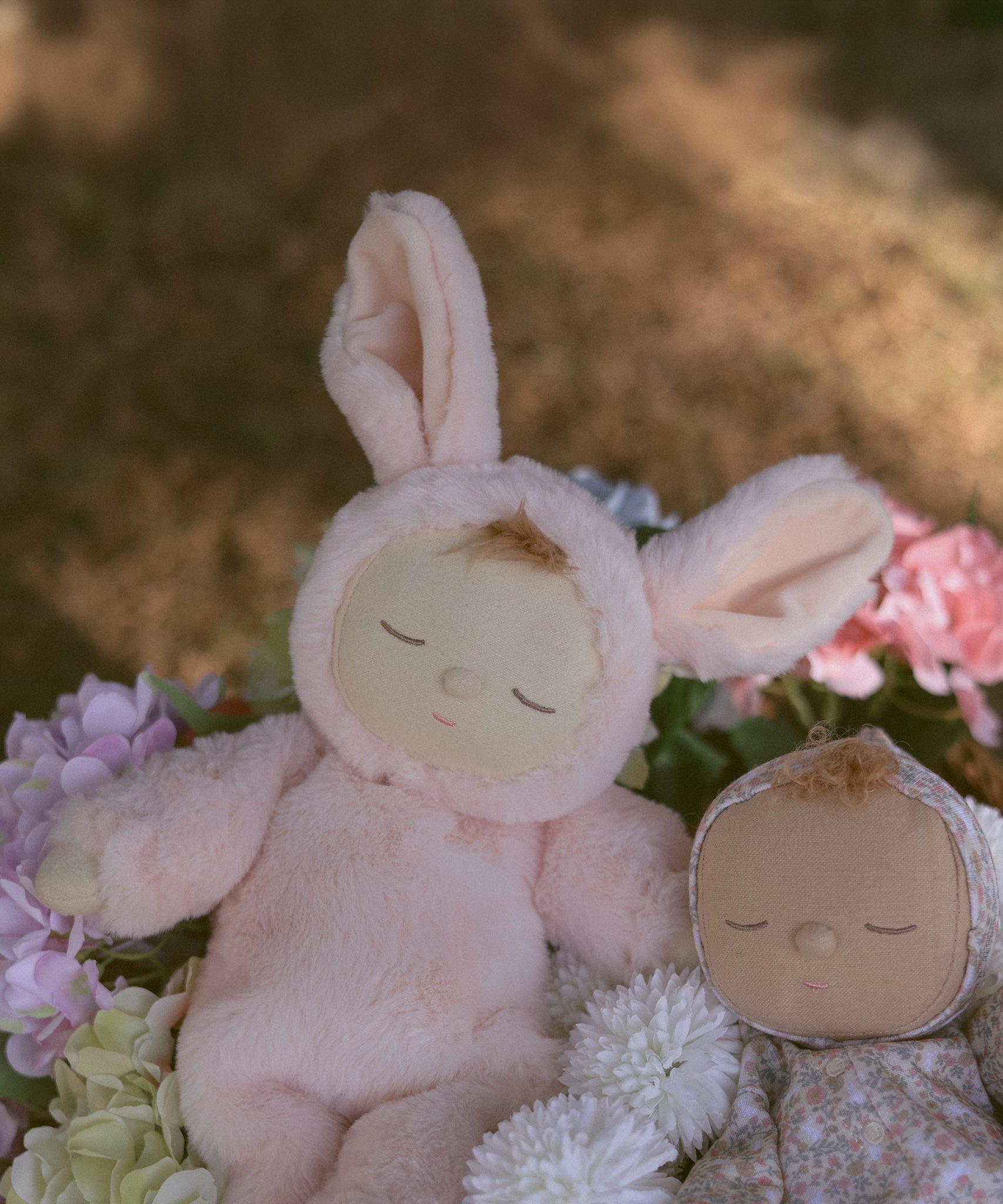 The Olli Ella Pink Bunny Moppet Lullaby Cozy Dinkum placed on a bunch of flowers with another dozy dinkum besides it. 