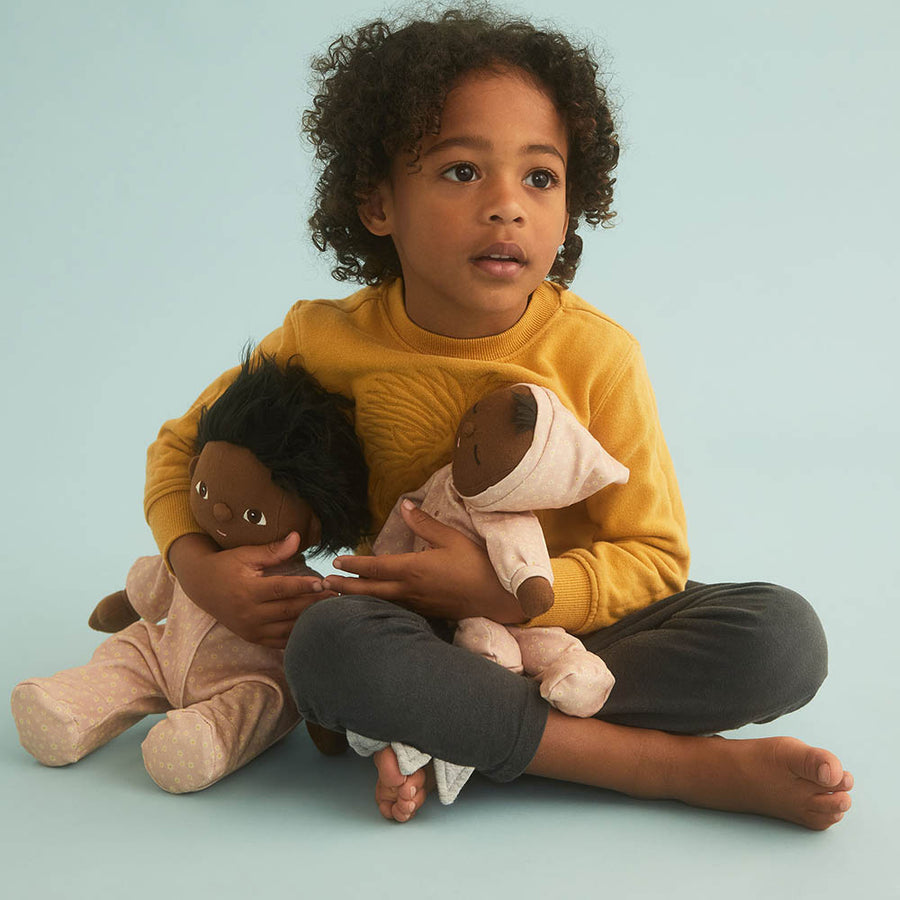 Young girl sat cross legged holding an Olli Ella mini daisy dozy dinkum and a dinkum doll on a blue background