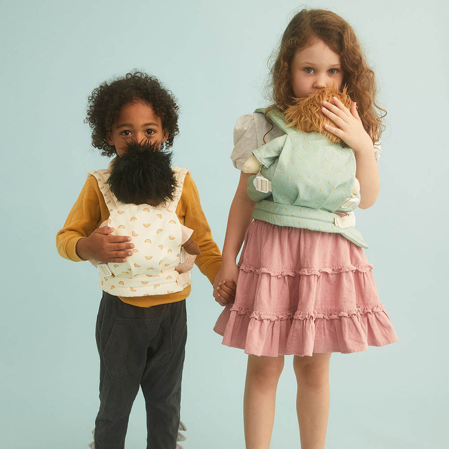 Two young children stood on a blue background holding Olli Ella dinkum dolls in toy doll baby carriers in the boat and rainbow print