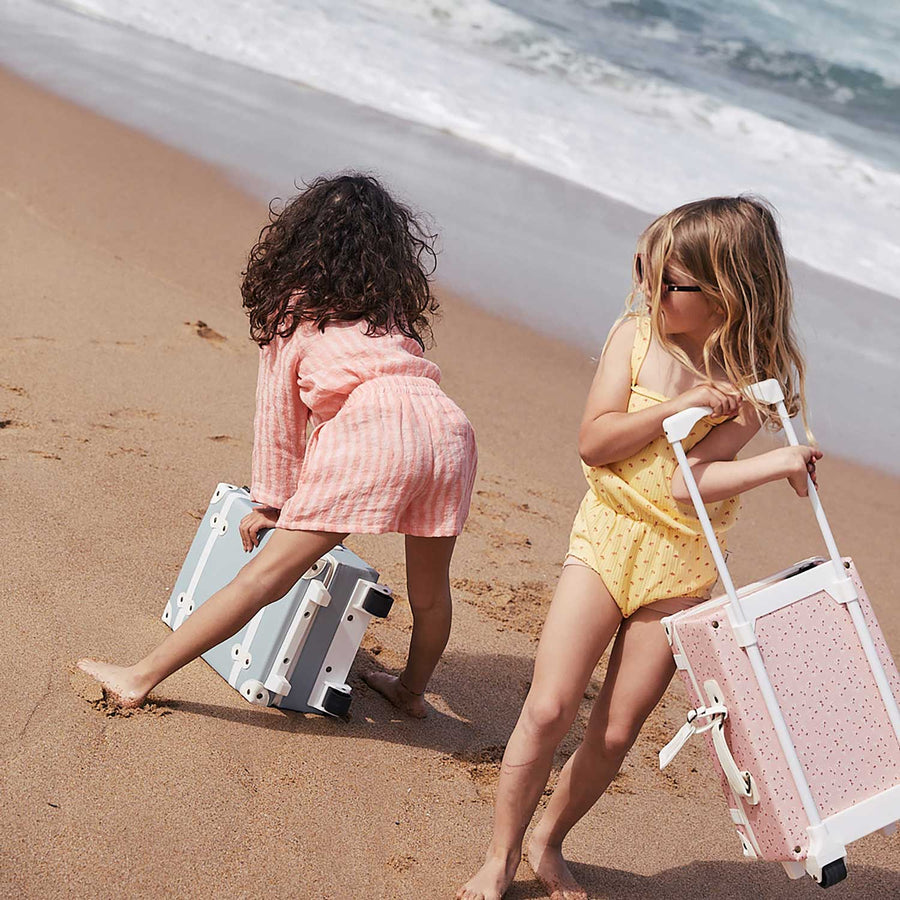 A child on a beach holding an Olli Ella Pink Daisies See-Ya Suitcase with another child next to them holding the leafed mushroom print one