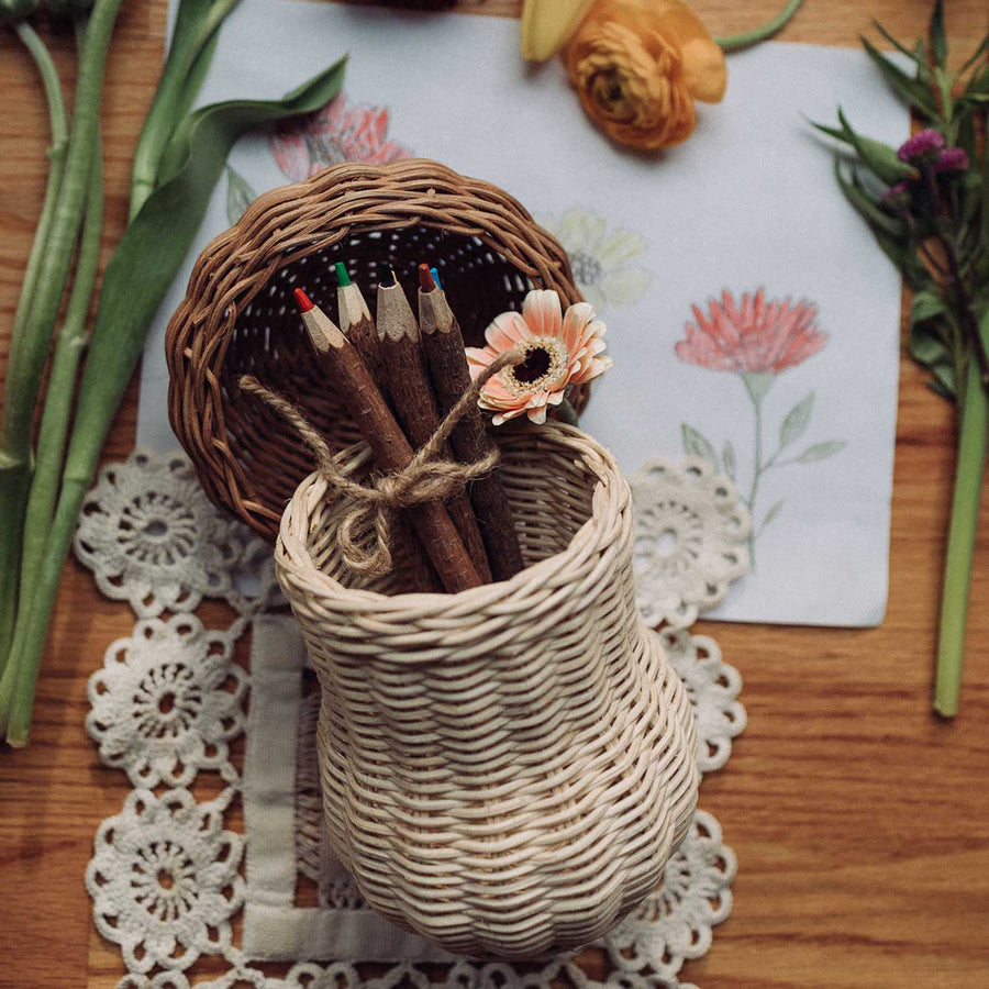 An Olli Ella Porcini basket shown on a table surrounded with flowers and a drawing of flowers. the lid is open and wooden twig pencils can be seen inside