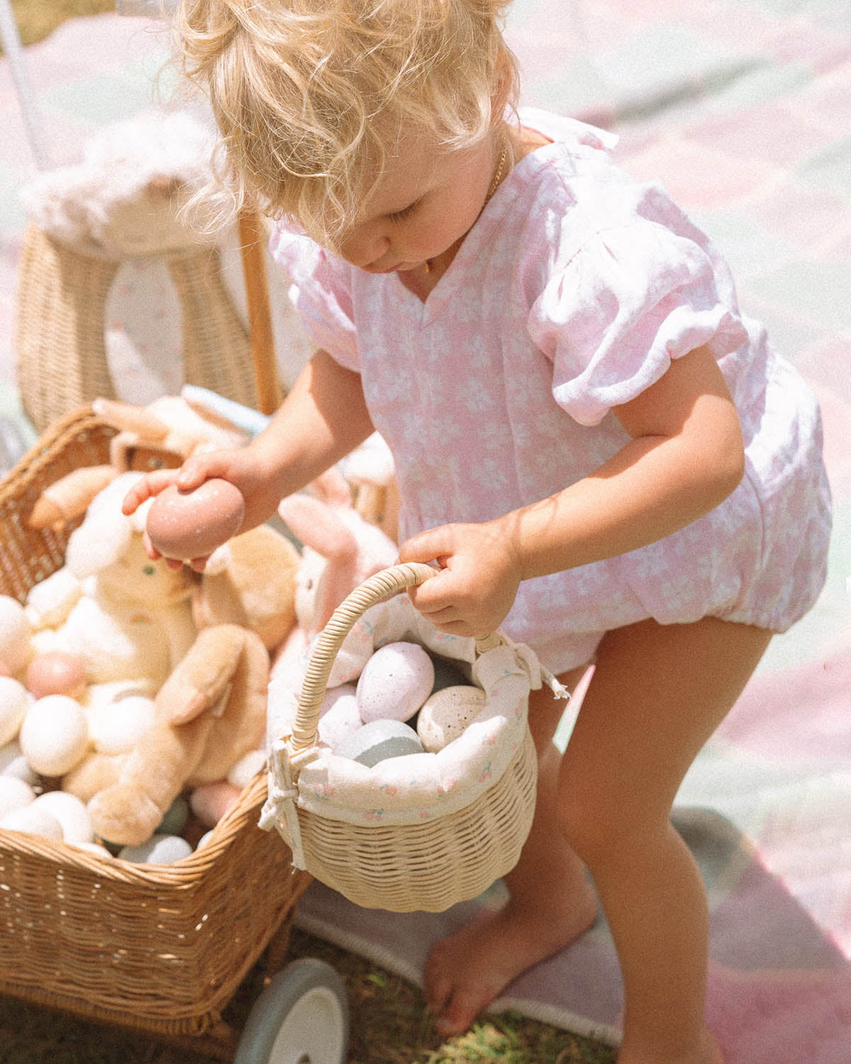 A child collecting eggs from a colourful blanket and into the Olli Ella Rattan Berry Basket with Lining – Pansy Floral.