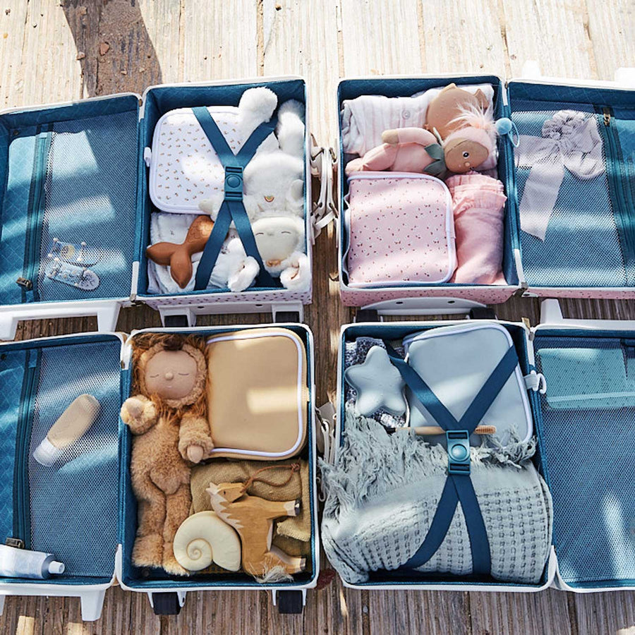 A child applying sun cream sitting next to an open olli ella see-ya suitcase filled with things including a matching butterscotch coloured wash bag