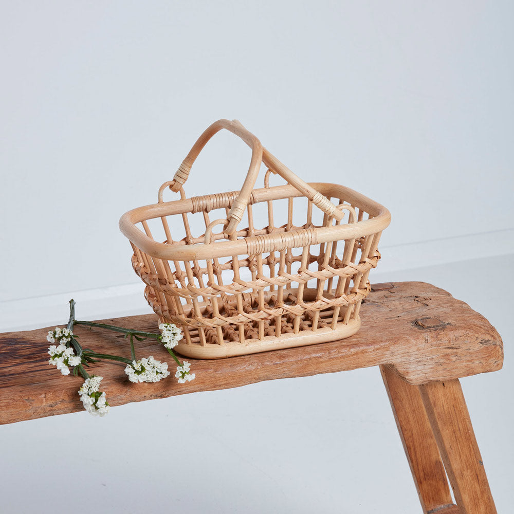 Olli ella eco-friendly woven rattan tarry basket in the wheat colour next to some white flowers on a wooden bench