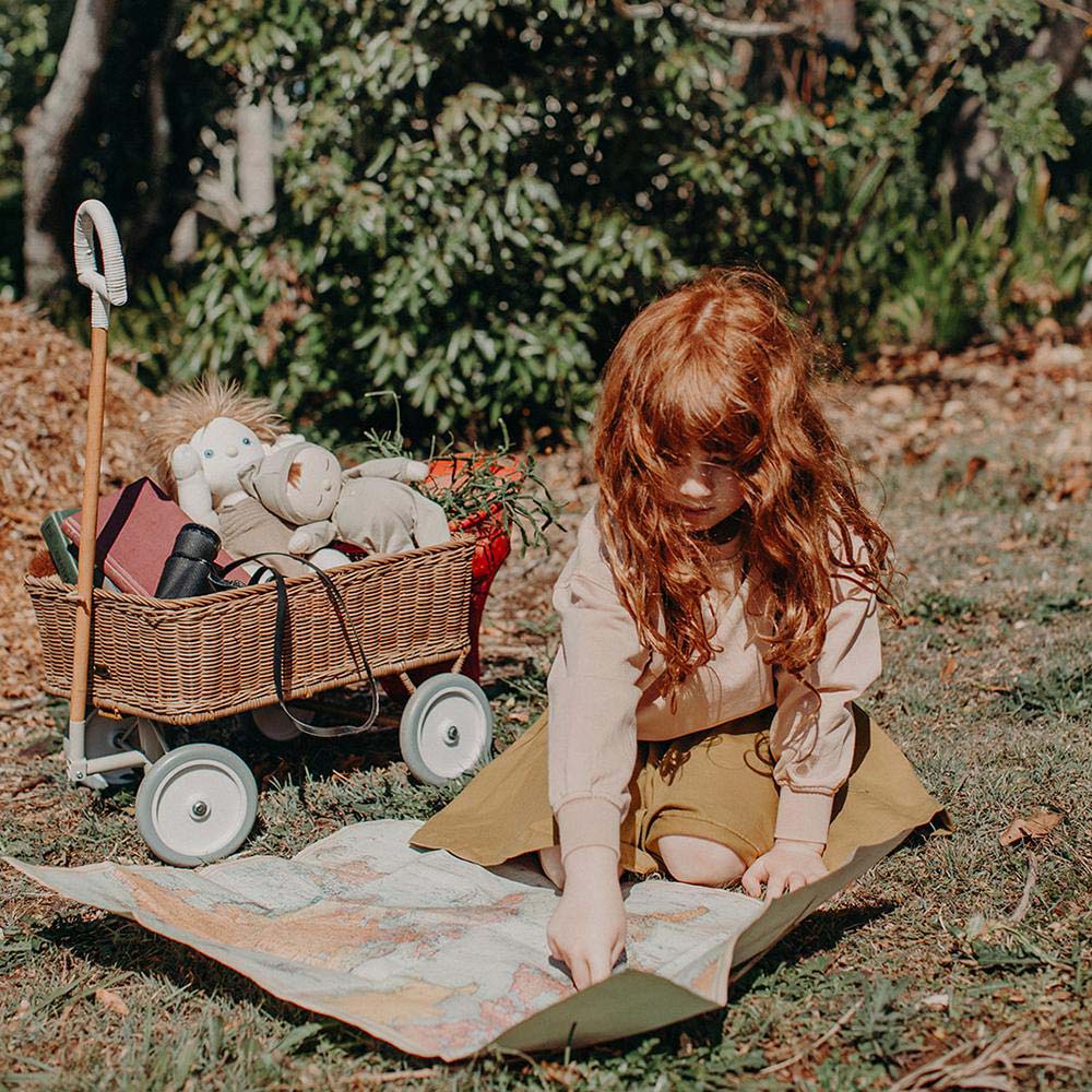 Girl looking at a large map on the floor next to an Olli Ella rattan wonder wooden wagon filled with toys