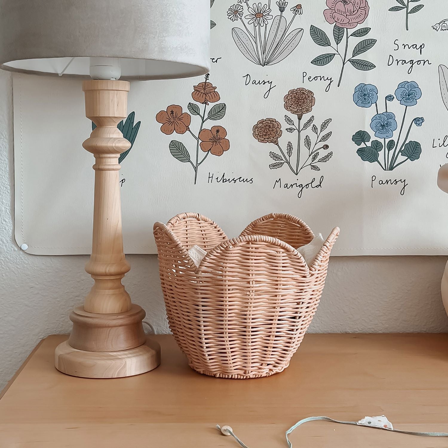 The Olli Ella Rattan Lily Basket Set in Seashell Pink on a side table next to a lamp with a flower poster on the wall in the background.