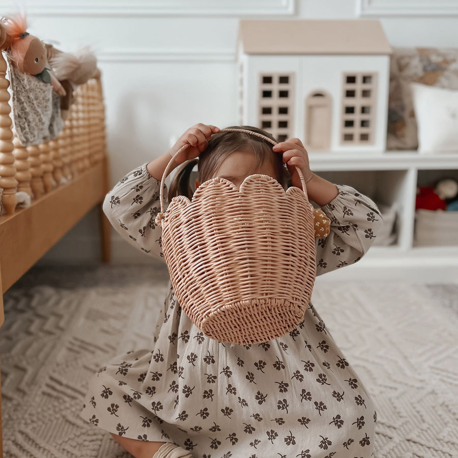 A young child wearing a white patterned dress in a nursery sat on the floor with the Olli Ella Rattan Tulip Carry Basket - Seashell Pink held up at head hight.