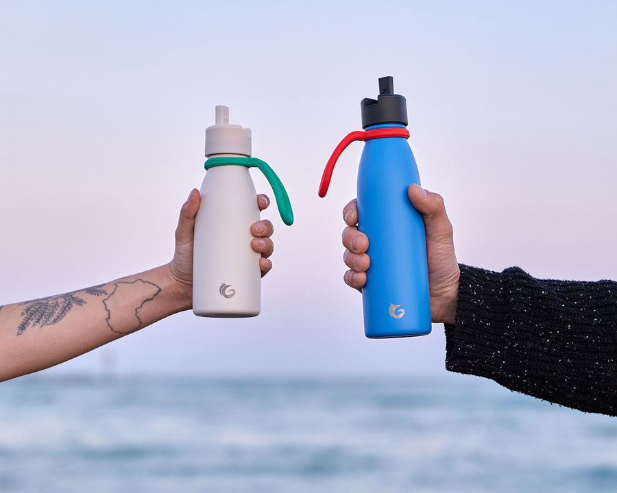 Two hands holding One Green Bottle canteens with Sloop bottle holders around the next in front of a sun set