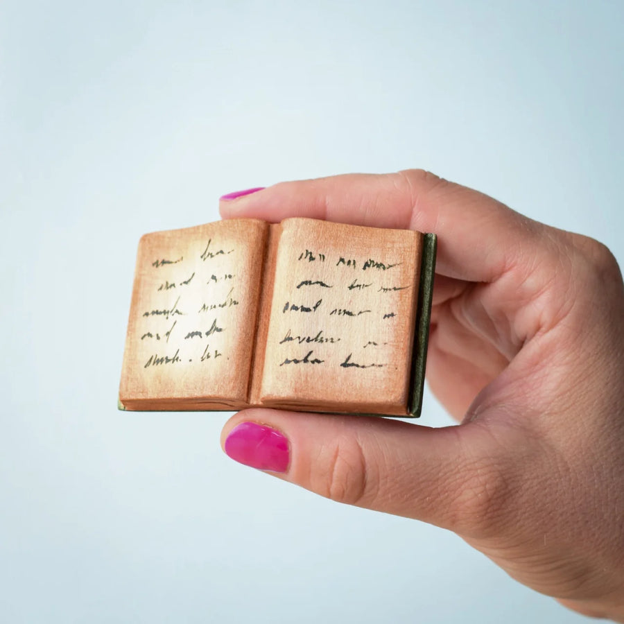 The Bumbu Toys Open Spell Book is being held in between a persons fingers, they are wearing pink nail polish. The pages in the book have been stained light brown to give it a vintage and old look